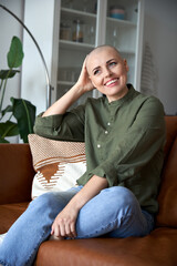 Happy bald woman breast cancer patient survivor sitting on couch at home. Smiling positive hairless lady touching shaved head feeling optimistic about recovery and remission relaxing at home. Vertical