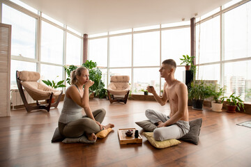 Young restful man and woman sitting on pillows and having tea on the floor