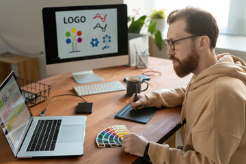 Young creative male freelance designer using tablet, stylus, laptop and palette