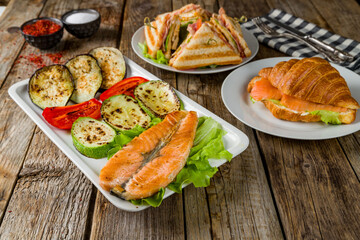 Fototapeta na wymiar Salmon steak on the grill, croissant with salmon and club sandwich on old wooden table