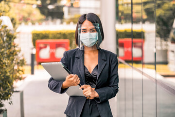 Asian young business woman walking and holding laptop wearing face shield with mask for healthcare which is required for doctor and general public to prevent COVID-19.