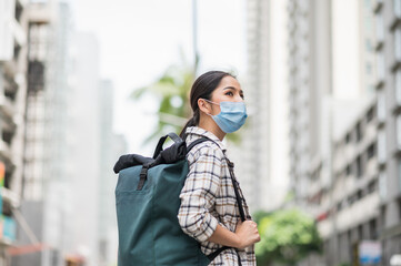 Fototapeta premium New normal of traveler. Side view of Alone young asian beautiful woman travel in holiday with backpack and wearing protection mask virus for safety trip. She walking alone in the city.