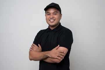 Smiling young handsome asian delivery man in black uniform standing with arm crossed on isolated...