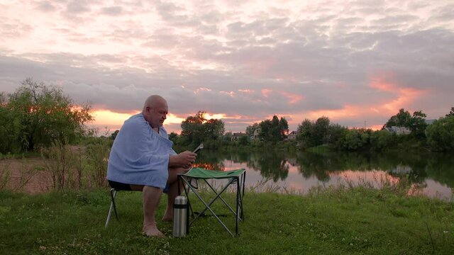 A young man is sitting with a towel on his shoulders. He holds the phone in his hand and dials the number. Rest on the shore of the lake. Against the background of the evening sky with a red sunset.