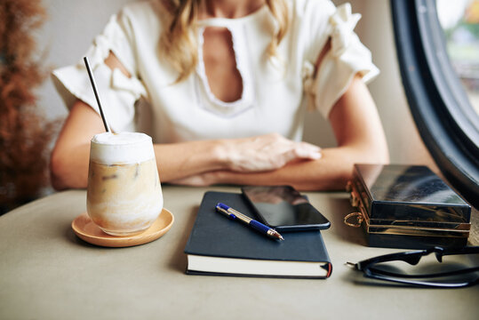 Cropped image of businesswoman sitting at cafe table with glass of iced coffee with smartphone and planner in front of her