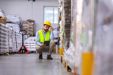 Young attractive bearded businessman in vest and helmet on head crouching and using tablet form...