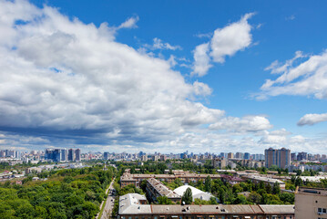 A cityscape with a green park in an old residential area of the city and new buildings on the horizon against a bright blue sky with thickening clouds.