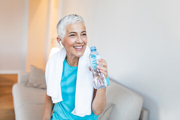 Elderly woman sitting on the sofa, exhausted after the daily training.Senior woman taking a break while exercising at home. Athletic mature woman holding bottle of water,Having a towel around her neck