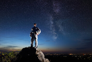 Space traveler standing on top of rocky hill and looking at beautiful sky with stars. Astronaut in...