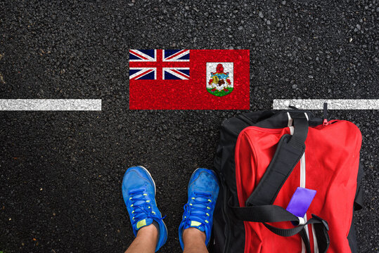 a man with a shoes and travel bag is standing on asphalt next to flag of Bermuda and border