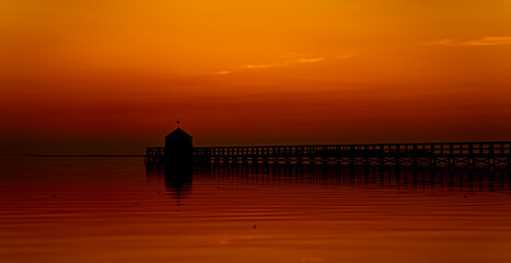 Naklejka na ściany i meble Large wooden pier on a beach just after sunset, bridge is in black silhouette and the sky of saturated with warm orange tones from the sunset.