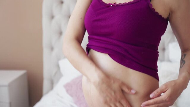 A Happy Pregnant Woman Applies Moisturizing Oil From Stretch Marks To Her Stomach Sitting At Home On The Bed, She Gently Strokes Her Tummy And Smiles. The Concept Of A Healthy Pregnancy