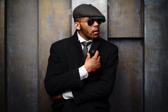 Studio shot of stylish african american man wearing coat, cap, glasses and holding smoking pipe