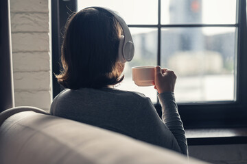 Back view of lonely сaucasian young woman enjoying having breakfast with cup of coffee and listenning music in headphones sitting near window at cold winter morning.