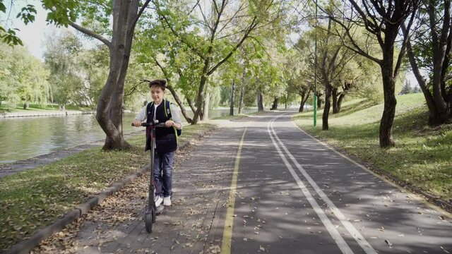 School boy riding with his electric scooter in the city with backpack on sunny day. Child in colorful clothes biking on way to school. Electric transport.