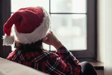 Back view of lonely sad young woman in a red santa claus christmas hat sitting and looking at window.