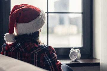 Back view of lonely sad young woman in a red santa claus christmas hat sitting near window and holding alarm clock. Concept