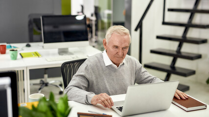 Portrait of aged man, senior intern looking focused at the screen while using laptop, sitting at...