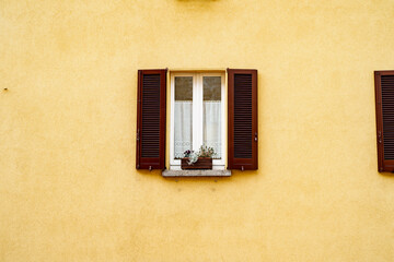 Fototapeta na wymiar A brown window with open shutters and flowers on the windowsill on a yellow wall.
