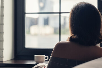 Back view of lonely сaucasian young woman wrapped in a plaid enjoying with cup of hot chocolate...