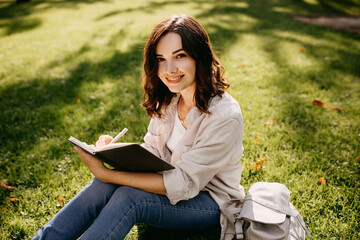 Obraz premium Young woman student sitting on grass in a park with notebook, writing, looking at camera.