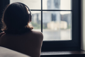 Back view of сaucasian young woman in headphones enjoying listening music sitting near window of apartments.