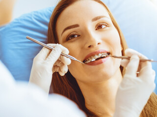 Smiling young woman with orthodontic brackets examined by dentist in sunny dental clinic. Healthy...