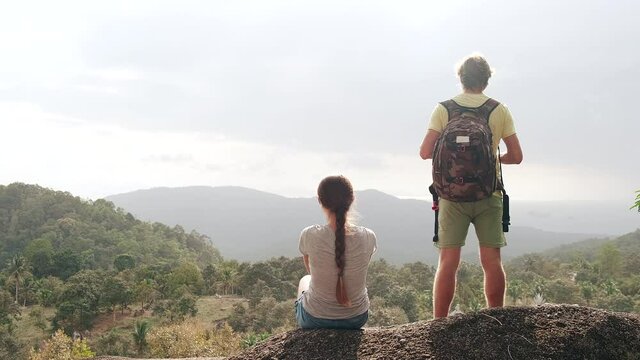 Rear view of two hikers with backpacks on the background of mountains. A man and a woman on a hill enjoy wild at sunset