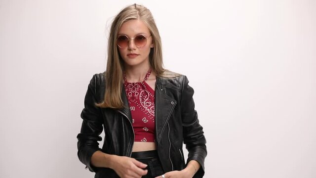 hippie young girl wearing sunglasses, holding hands in pockets, arranging jacket and sunglasses, turning and smiling, laughing on grey background