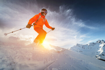 A man freerider skier rides at speed in snow powder from a slope against a background of snow-capped mountains in the rays of the sunset