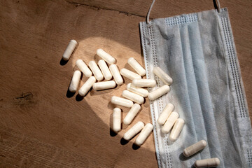 A handful of white pills are scattered on the table next to the medical mask. White medical capsule tablets