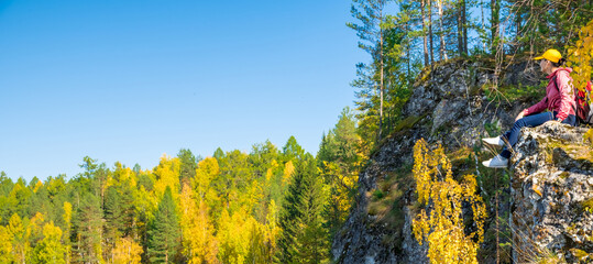 Girl tourist with a backpack looks at the autumn forest while sitting on a rock, Ural, Russia. Panoramic banner