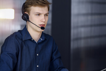 Young blond businessman using headset and computer in a darkened office, glare of light on the background. Startup business means working hard