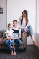 Family photosession in studio. Little daughter mom and dad sits on sofa by the window