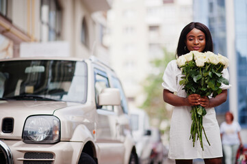 Fototapeta premium Beautiful african american girl holding bouquet of white roses flowers on dating in the city. Black businesswoman with bunch of flowers near car.