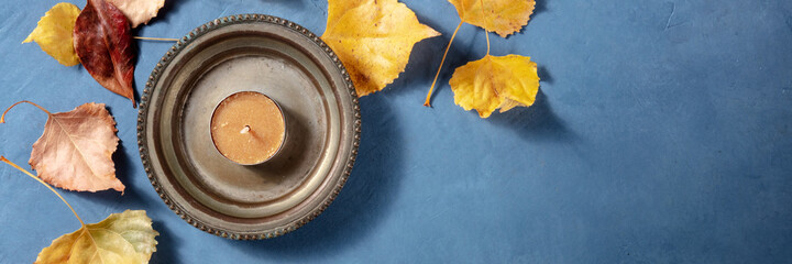 Autumn panoramic banner with yellow leaves, a candle, and copyspace, a flat lay on a blue background