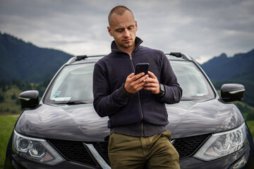 A young man is leaning against his broken car and is writing a report to the insurance company.