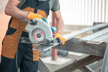 A carpenter in overalls saws a black-painted Board with a circular saw.