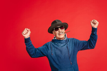 Winner, smoking. Portrait of senior man in stylish eyewear and hat isolated on red studio background. Tech and joyful elderly lifestyle concept. Trendy colors, forever youth. Copyspace for your ad.
