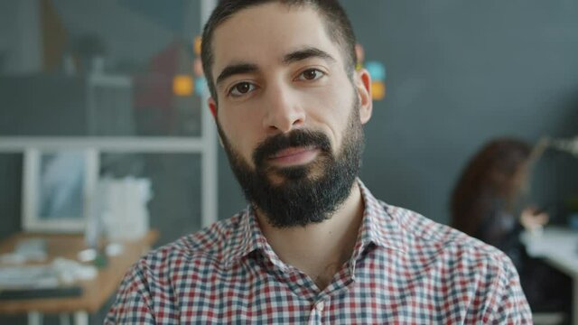 Slow motion portrait of handsome Arab guy looking at camera in shared office, woman is working in background. People, business and emotions concept.