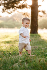 Happy adorable baby boy walks on the grass in the park on summer day. Happy smile. Child in trendy and cute clothes