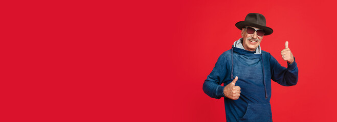 Thumbs up. Portrait of senior man in stylish eyewear and hat isolated on red studio background. Tech and joyful elderly lifestyle concept. Trendy colors, forever youth. Copyspace, flyer horizontal