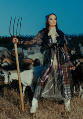 Woman farmer stands with pitchfork among a herd of goats.