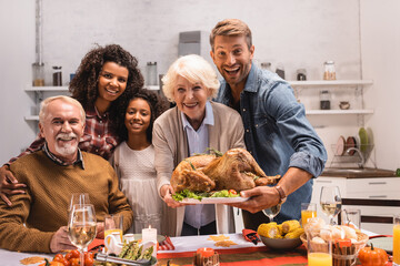Selective focus of multiethnic family holding turkey and looking at camera during thanksgiving