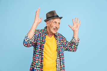Shocked, astonished. Portrait of senior hipster man in stylish hat isolated on blue studio background. Tech and joyful elderly lifestyle concept. Trendy colors, forever youth. Copyspace for ad.