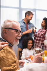 Fototapeta premium Selective focus of elderly man holding glass of wine while celebrating thanksgiving with multicultural family at home