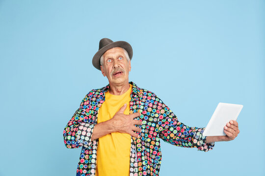 Videochat using tablet, shocked. Portrait of senior hipster man in stylish hat on blue studio background. Tech and joyful elderly lifestyle concept. Trendy colors, forever youth. Copyspace for ad.