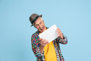 Playing games using tablet. Portrait of senior hipster man in stylish hat isolated on blue studio...