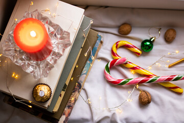 Christmas or New Year's comfort. Composition with a stack of books, a red candle, seasonal sweets, Christmas tree decorations and lights. Home atmosphere, relaxation, winter holidays