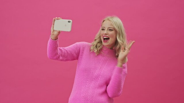 A smiling blonde woman in a pink sweater is taking selfie photos using her smartphone standing isolated over a pink background